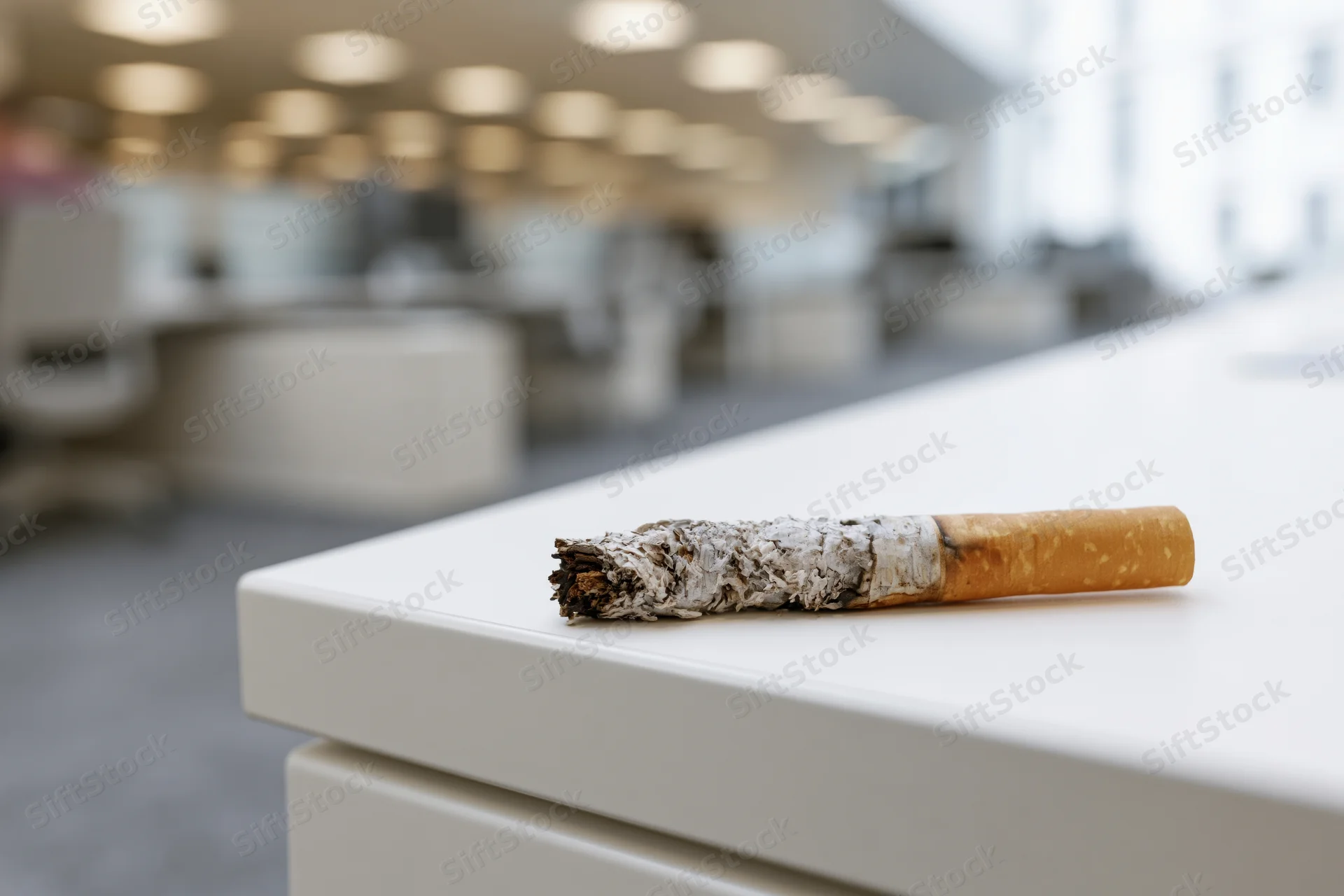 Burnt cigarette resting on white office desk with blurred workspace background