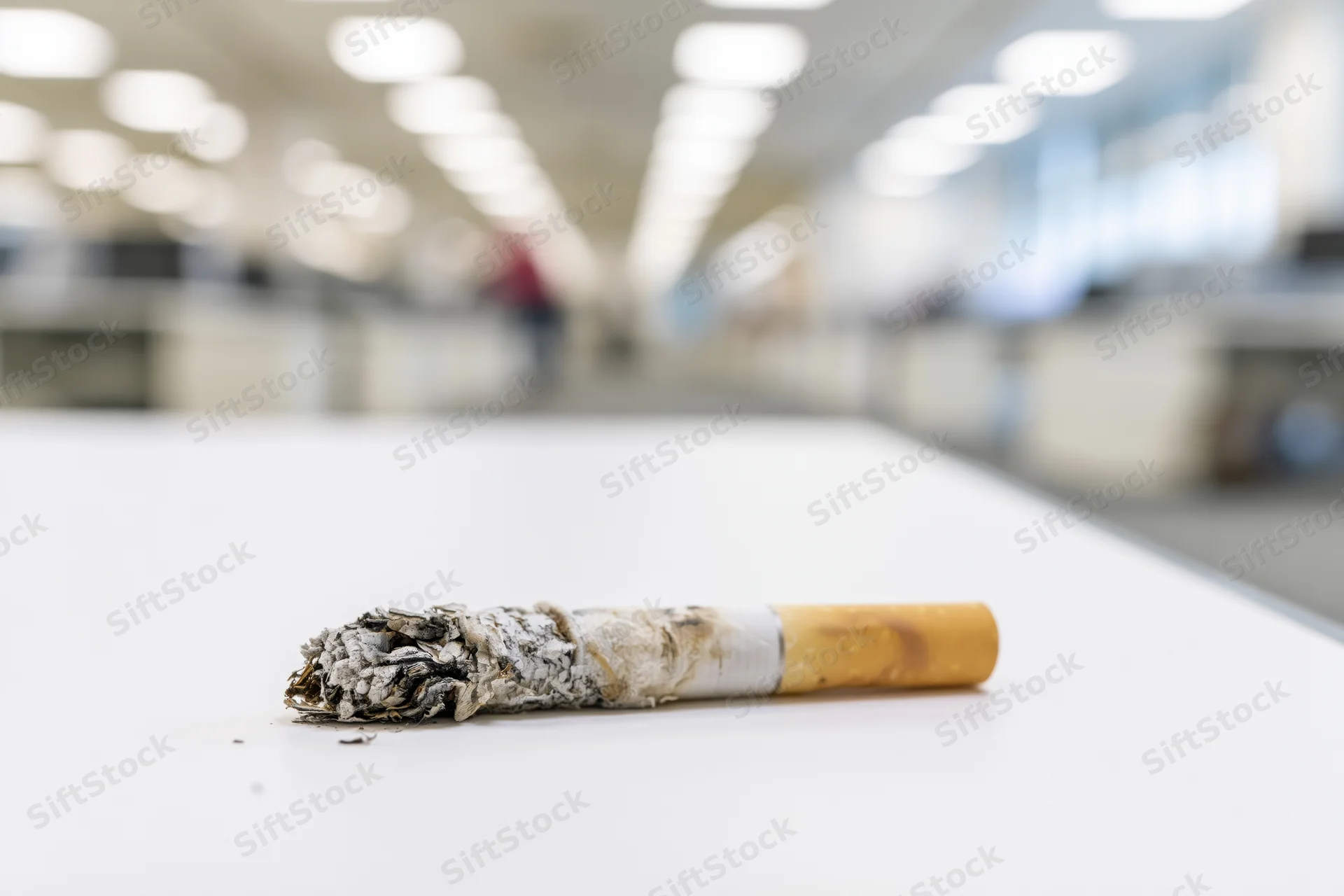 Burnt cigarette with ash resting on white office desk in blurred workplace setting