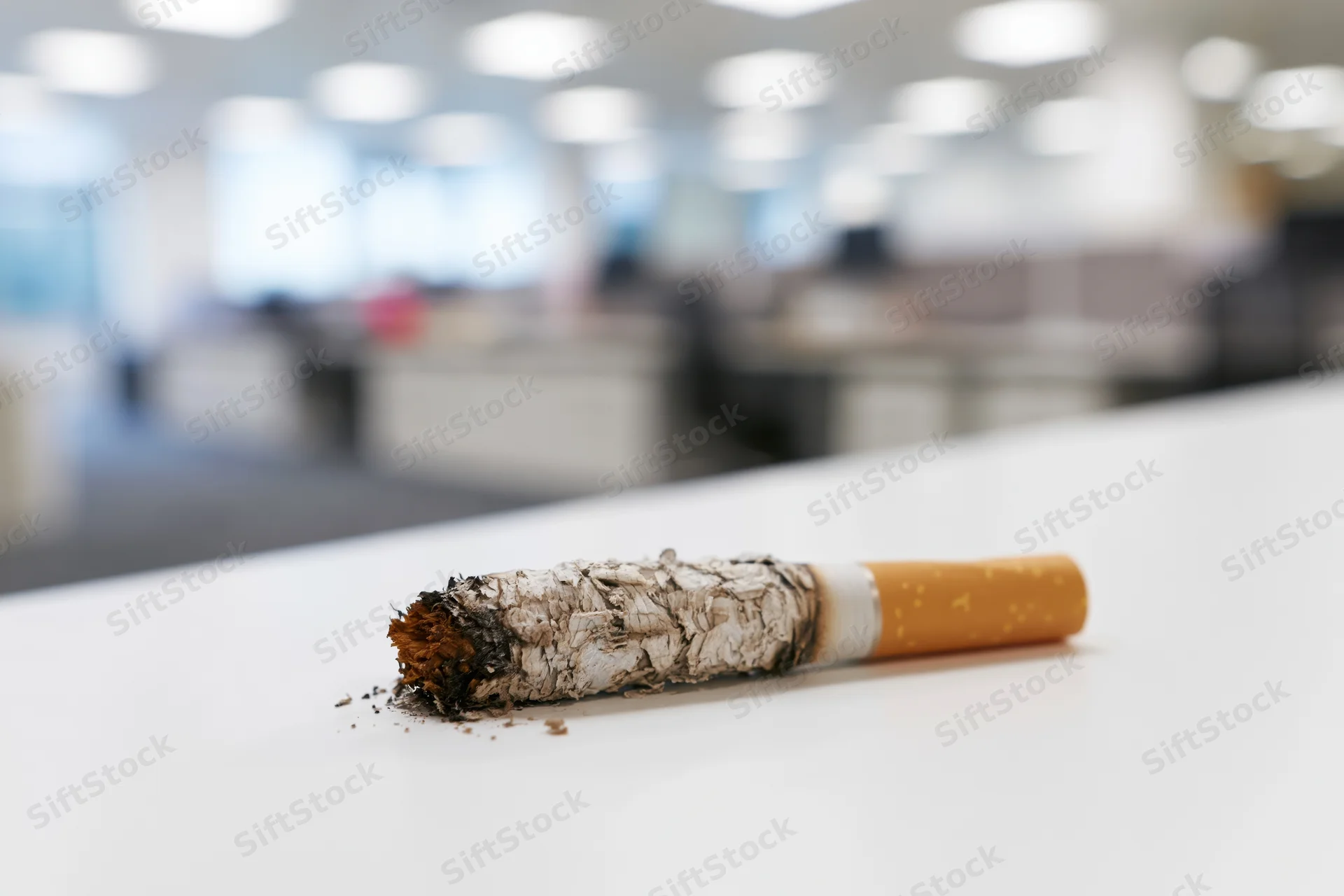 Burnt cigarette with ash resting on white office desk, blurred workspace in background