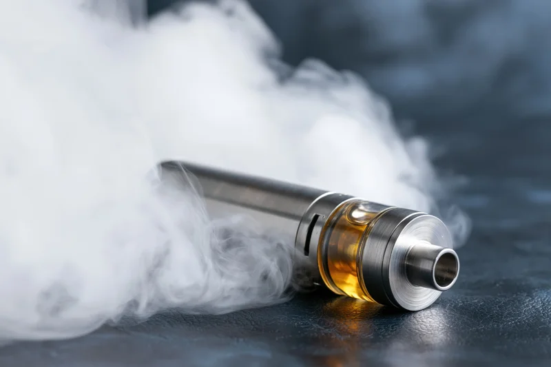 Cylindrical vape mod lying on dark textured surface with white vapor swirling around it