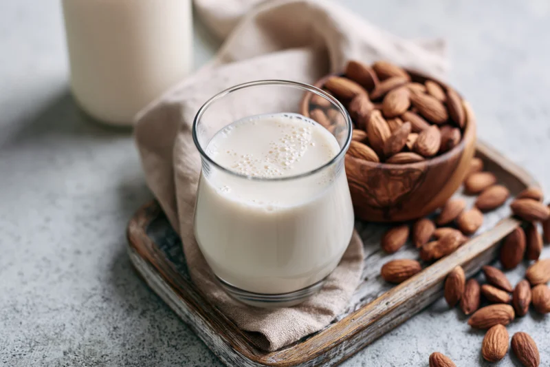 Glass of almond milk with foam on a wooden tray beside a bowl of almonds and a cloth napkin
