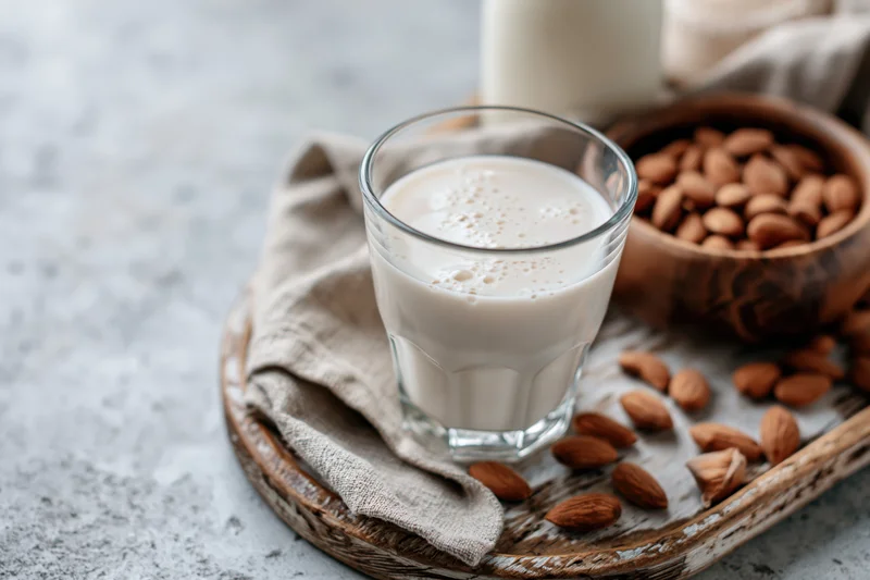 Glass of almond milk with foam on a wooden tray beside a bowl of almonds and a cloth napkin