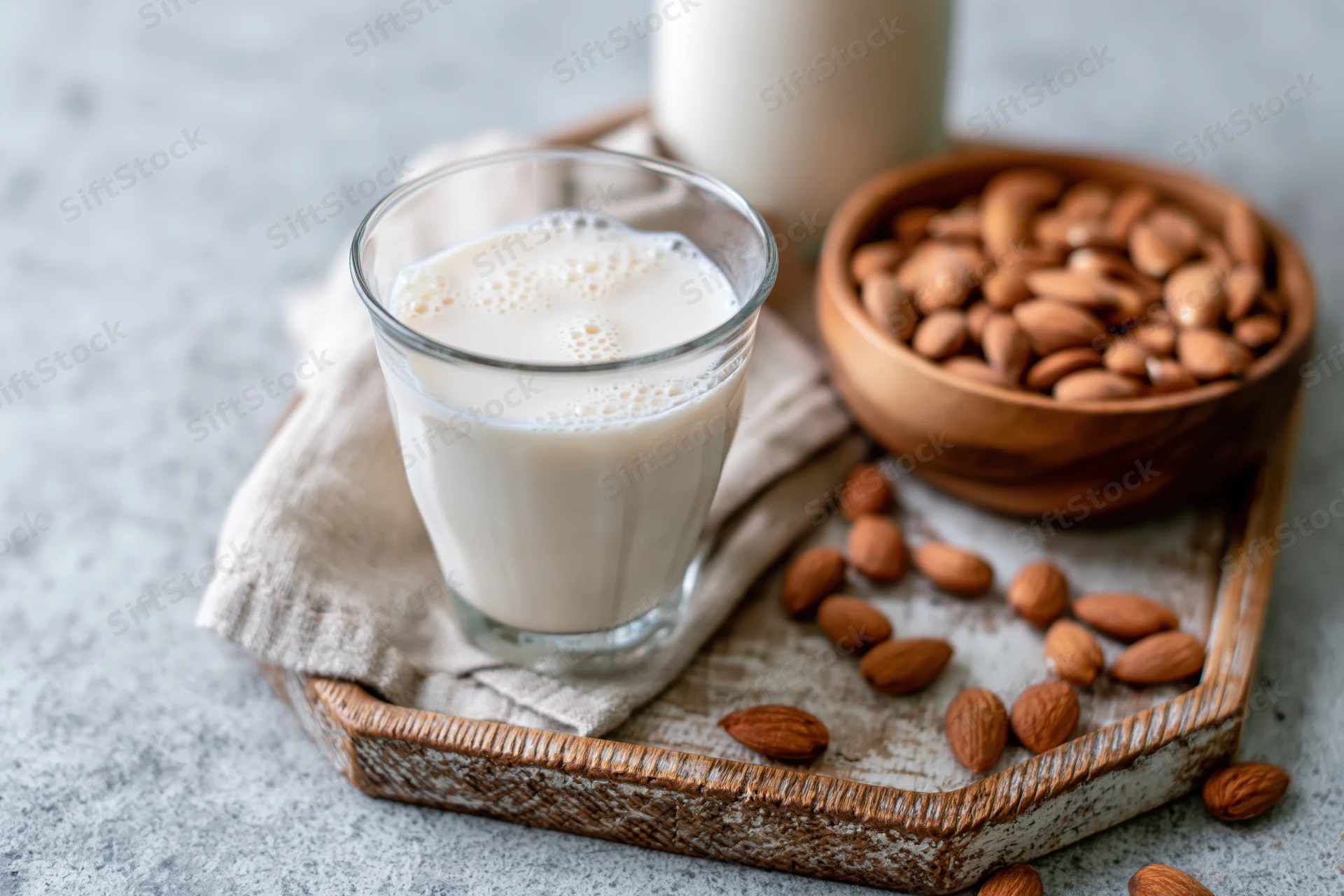 Glass of almond milk with foam on a wooden tray beside a bowl of almonds and scattered nuts on a light countertop