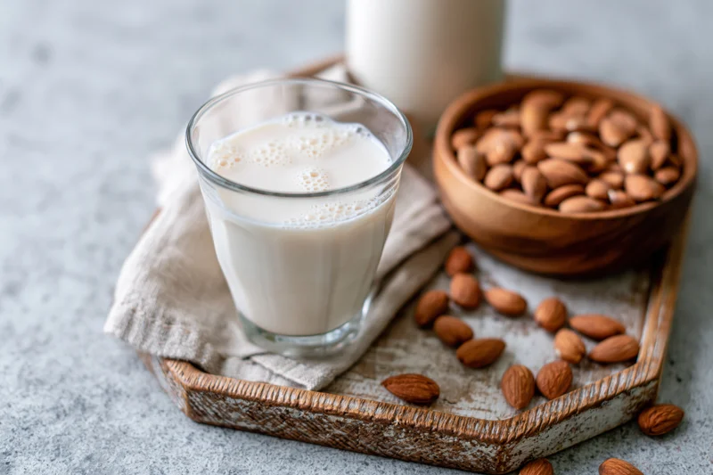 Glass of almond milk with foam on a wooden tray beside a bowl of almonds and scattered nuts on a light countertop