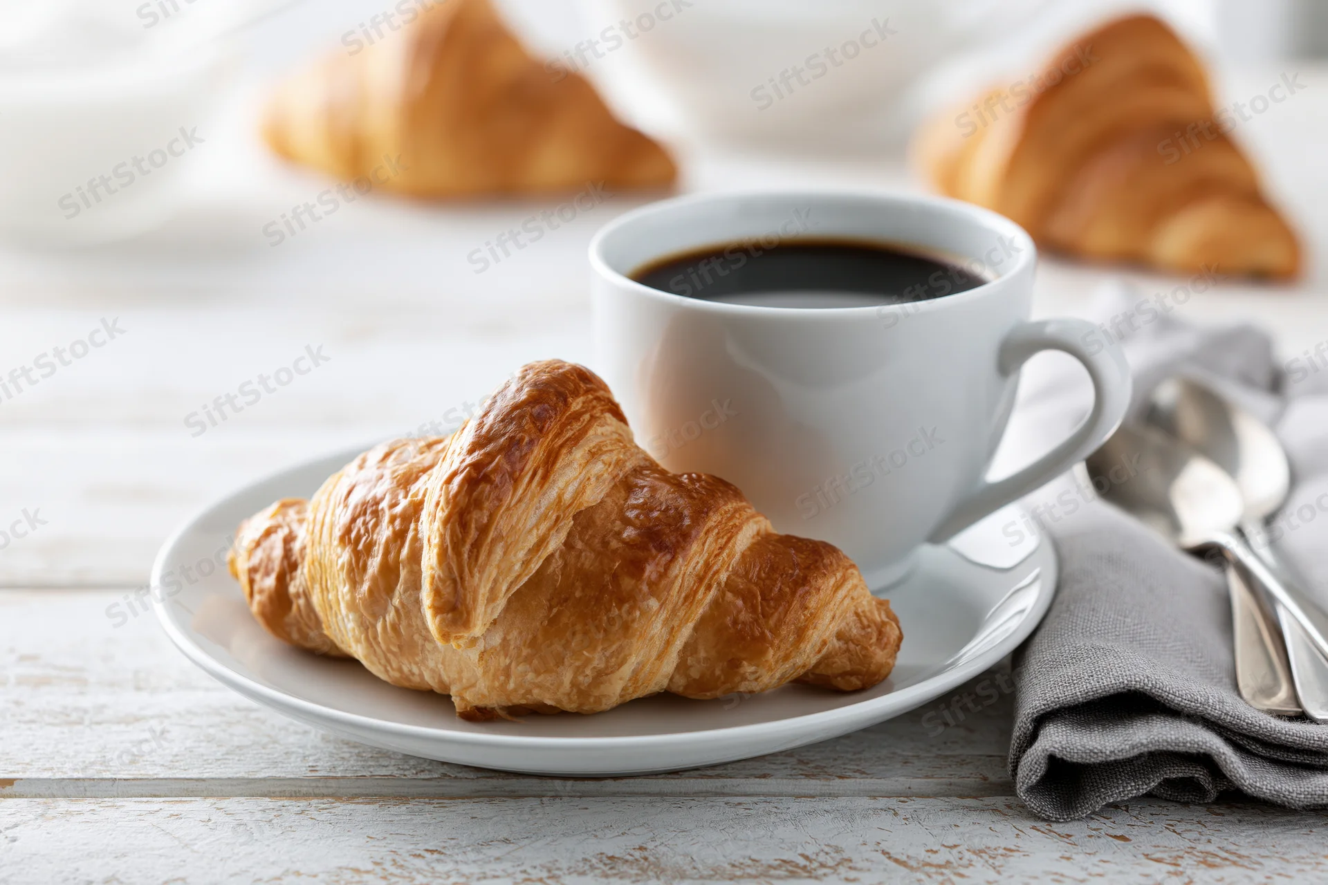 Golden-brown croissant on a white saucer with a cup of black coffee on a wooden table