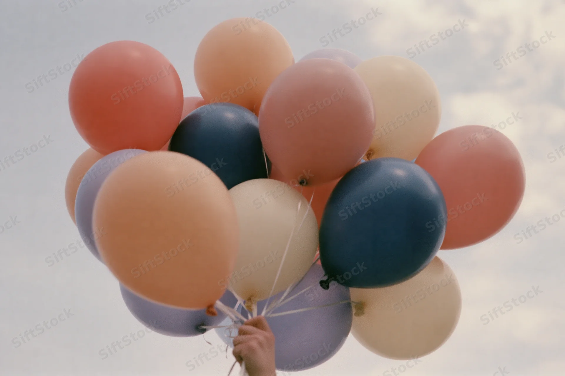 Hand holding a cluster of pastel-colored helium balloons against a cloudy sky