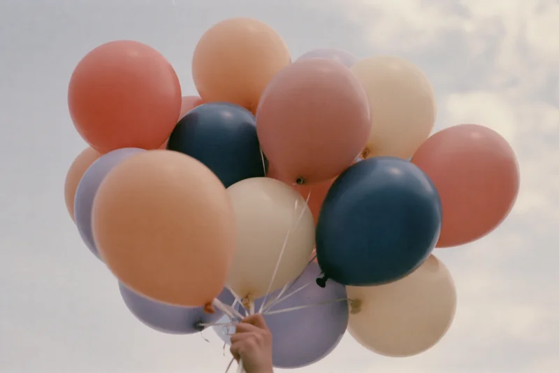 Hand holding a cluster of pastel-colored helium balloons against a cloudy sky