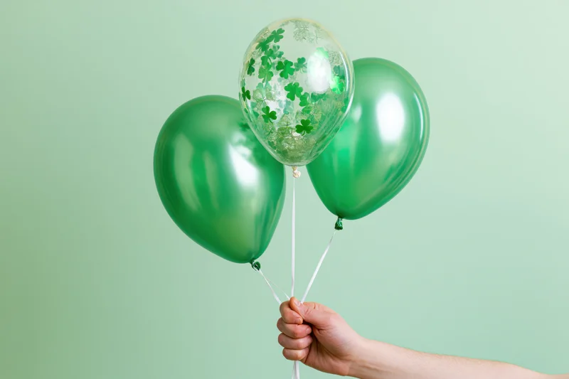 Hand holding three green balloons with shamrock pattern against light green background