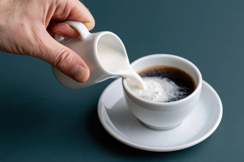 Hand pouring milk into a cup of coffee on a saucer against a dark teal surface