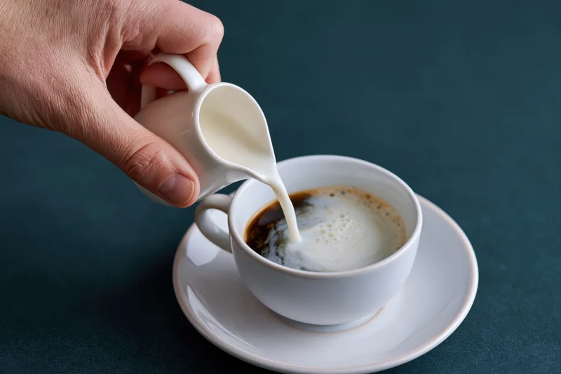 Hand pouring steamed milk into a white coffee cup on a saucer