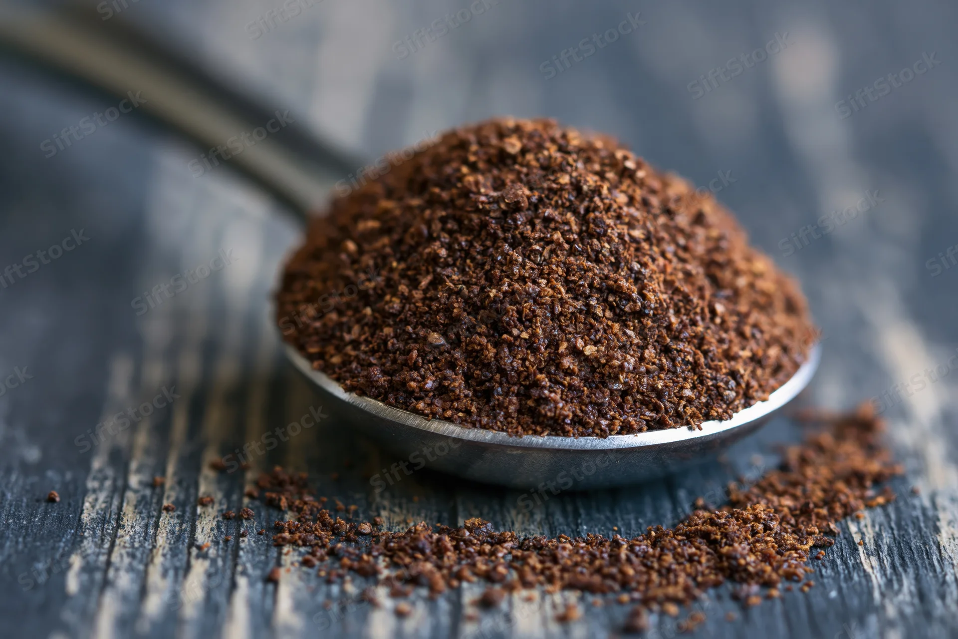 Heaping metal measuring spoon filled with coarse ground coffee resting on dark wooden surface