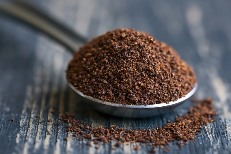 Heaping metal measuring spoon filled with coarse ground coffee resting on dark wooden surface