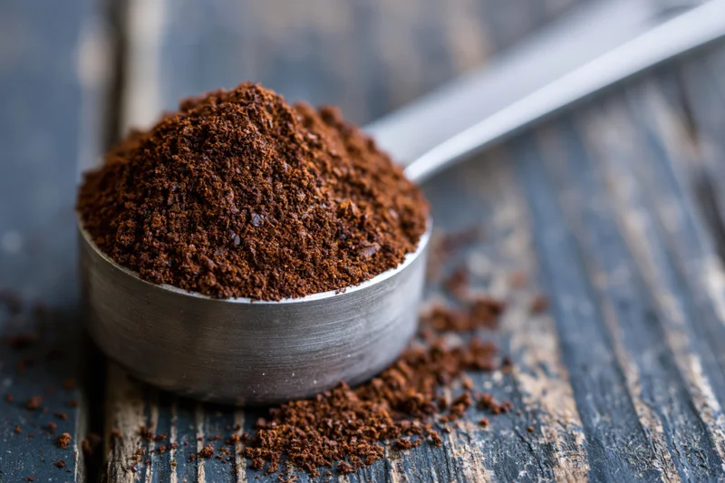 Heaping metal measuring spoon filled with coarse ground coffee resting on a dark wooden surface