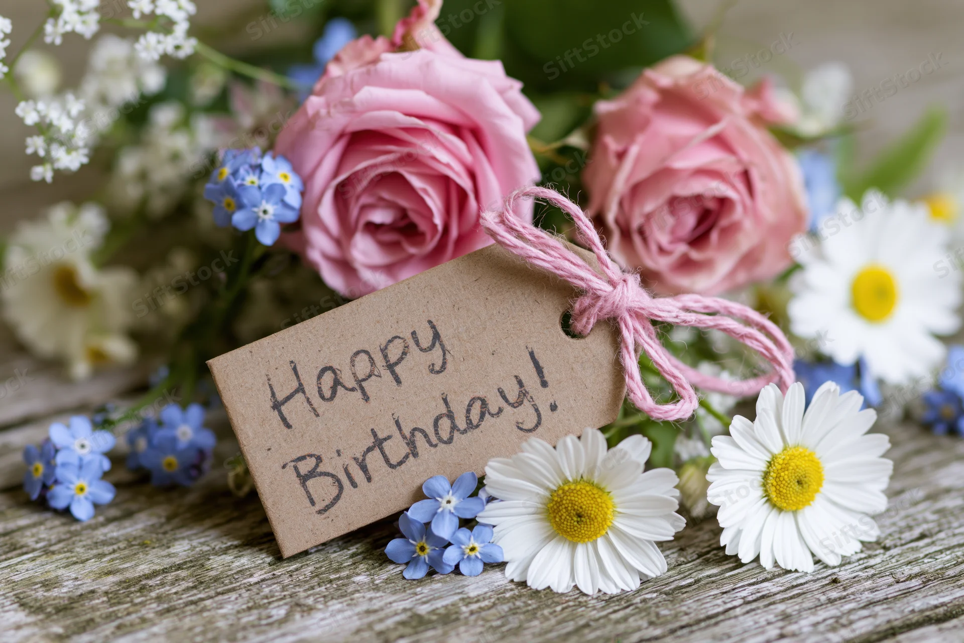 Pink roses and white daisies with a kraft paper tag reading happy birthday tied with pink string on a wooden table