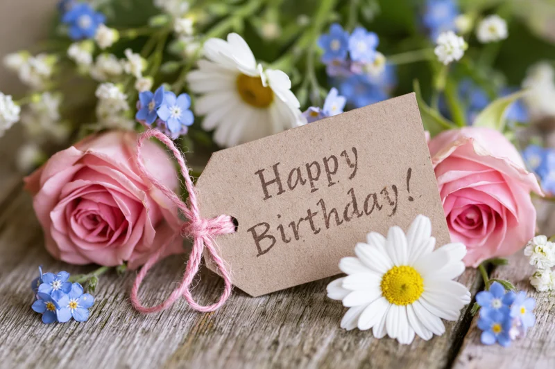 Kraft paper gift tag reading Happy Birthday tied with pink string among pink roses, white daisies and blue forget-me-nots on a wooden surface