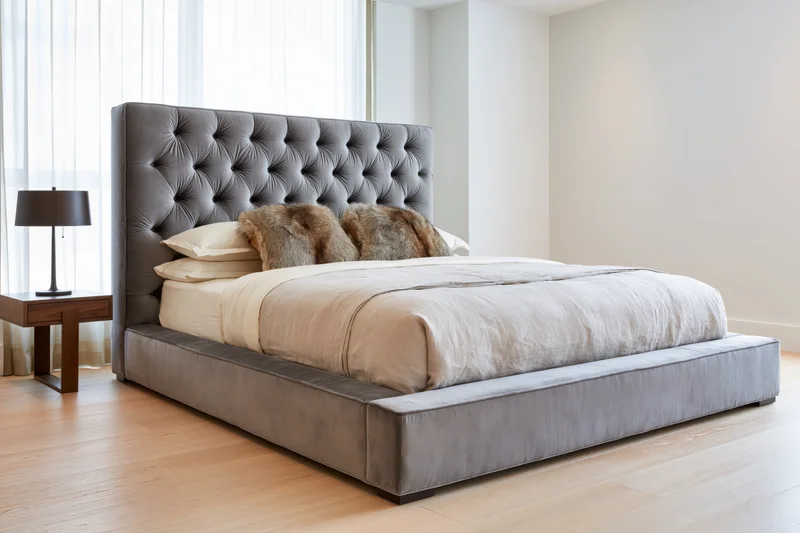 Large upholstered platform bed with tall tufted velvet headboard in modern bedroom with wooden flooring and natural light