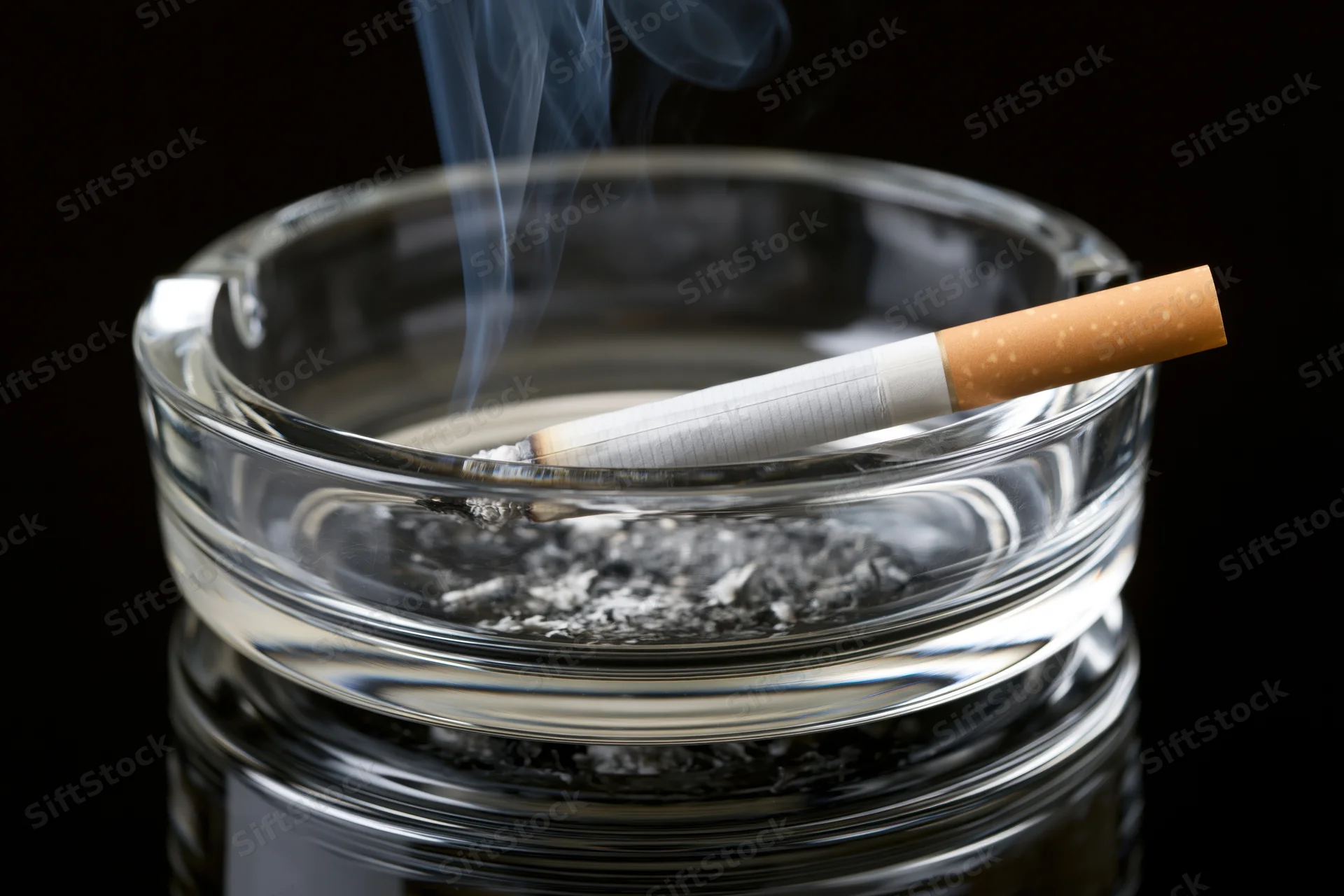 Lit cigarette resting in clear glass ashtray with smoke rising against black background