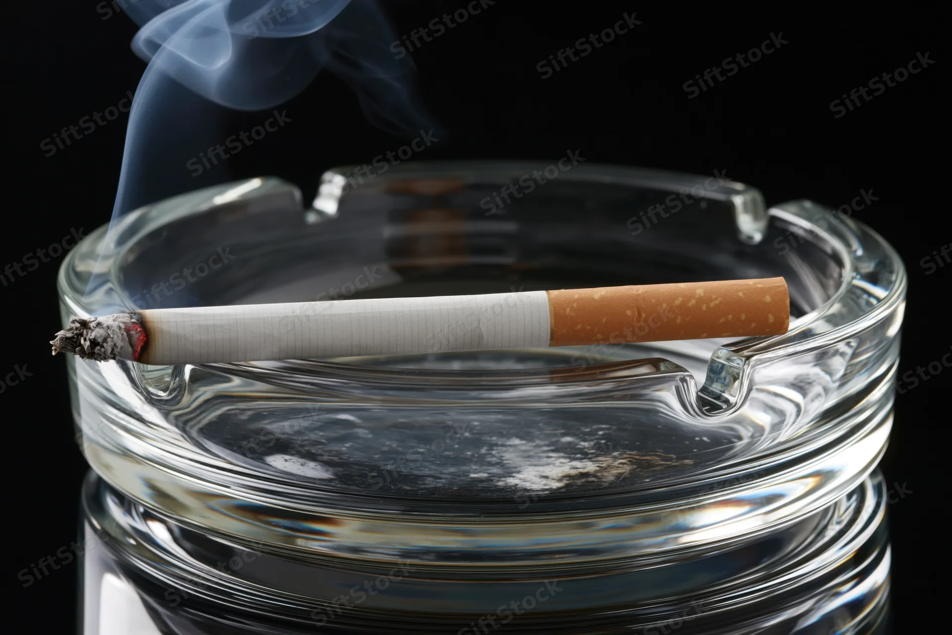 Lit cigarette resting in clear glass ashtray with smoke rising against black background