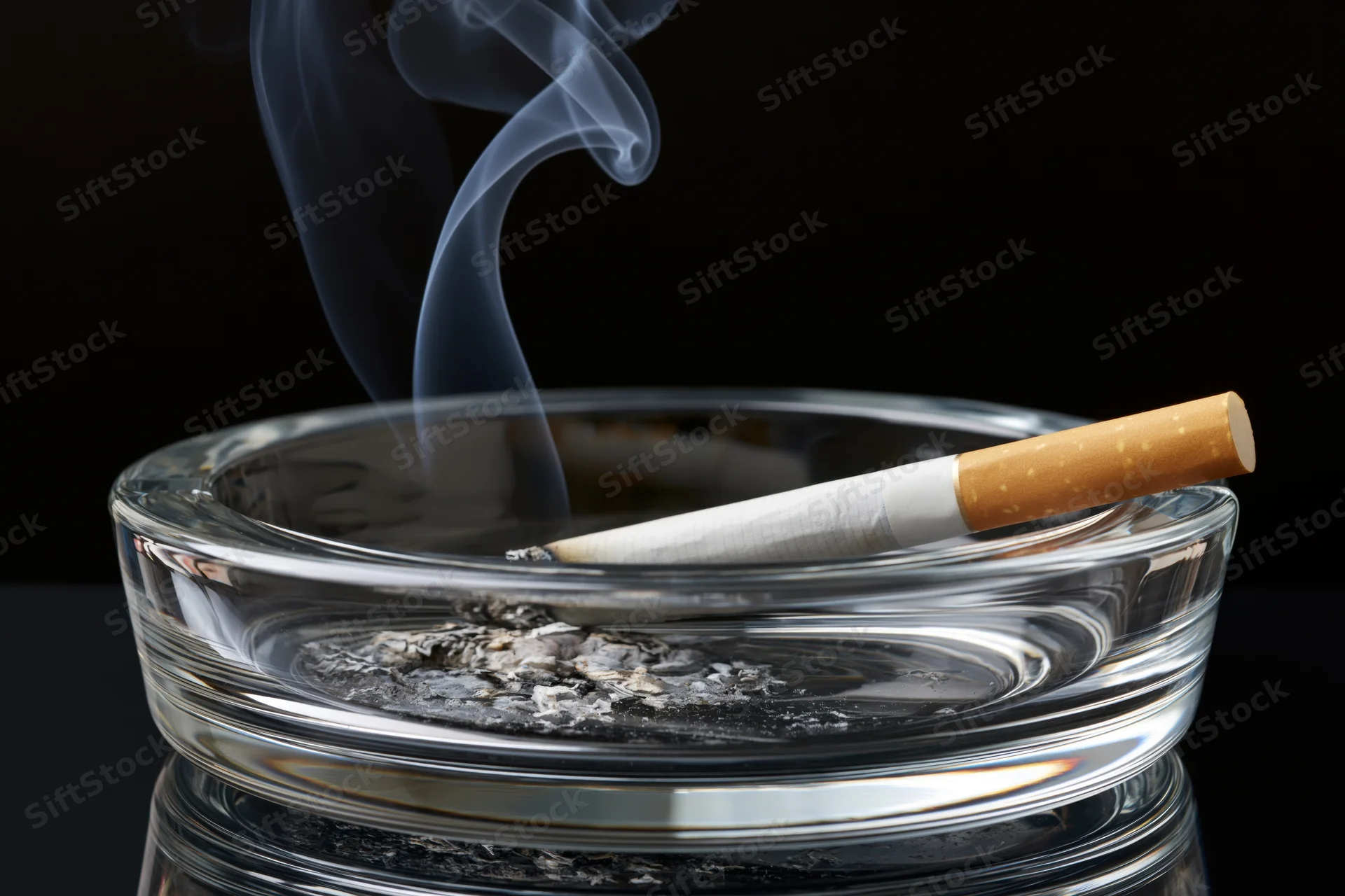 Lit cigarette resting in a clear glass ashtray with smoke rising against black background