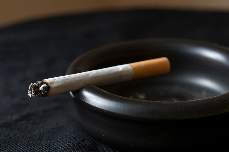 Lit cigarette with ash resting in notch of black ceramic ashtray on dark fabric surface