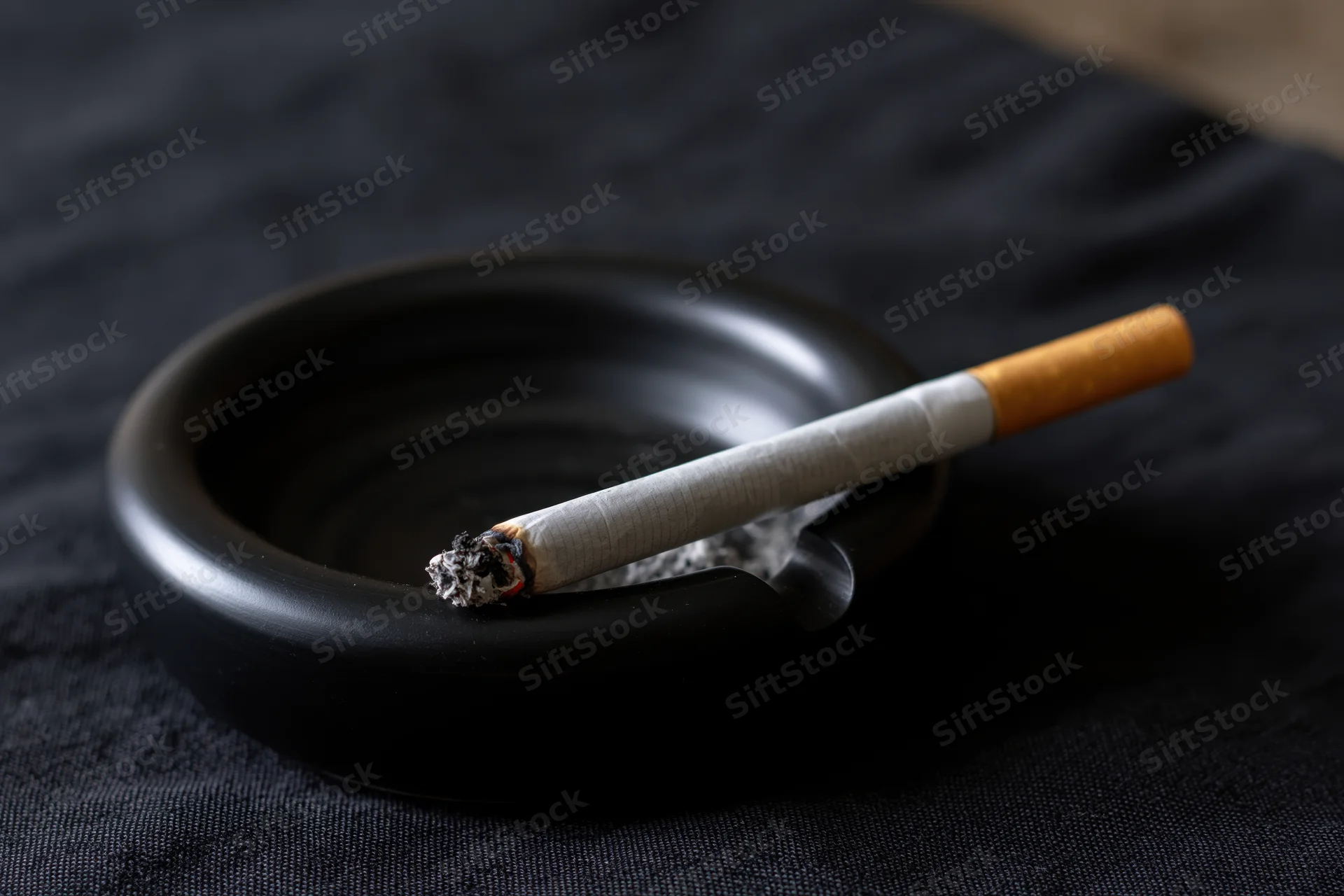 Lit cigarette resting in notch of black ceramic ashtray on dark fabric