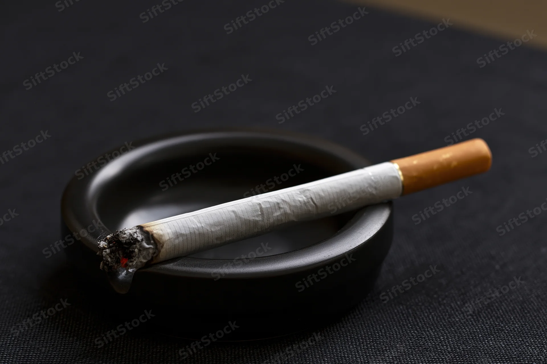 Lit cigarette resting in notch of small black ceramic ashtray on dark textured surface