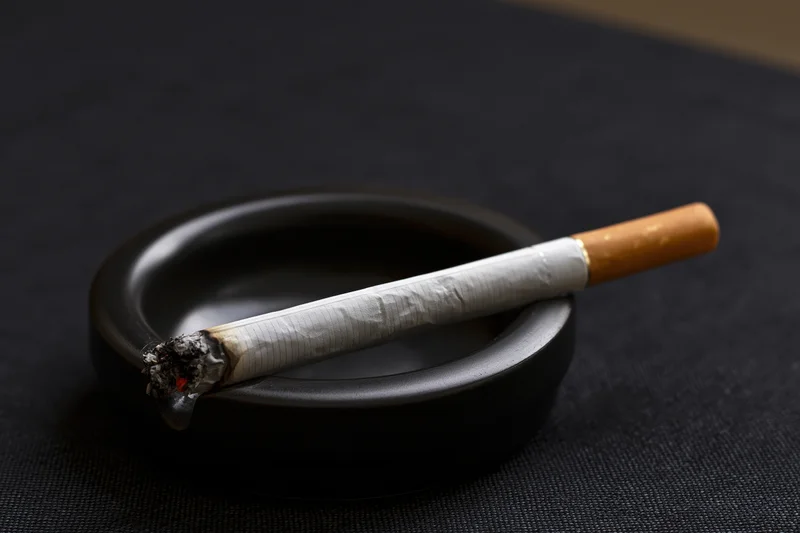 Lit cigarette resting in notch of small black ceramic ashtray on dark textured surface