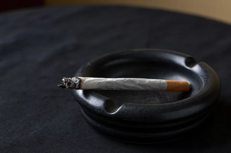 Lit cigarette resting in notch of black ceramic ashtray on dark surface