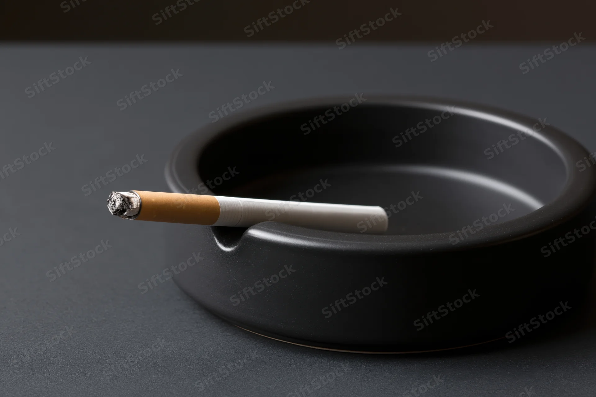Lit cigarette resting in notch of black round ashtray on dark surface
