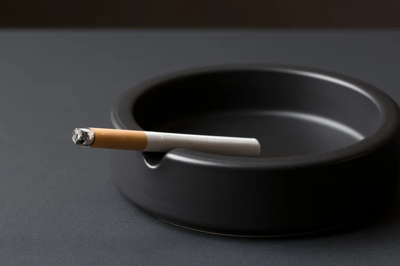 Lit cigarette resting in notch of black round ashtray on dark surface