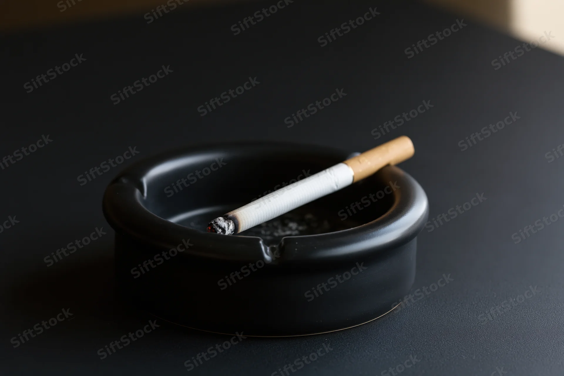 Lit cigarette resting in notch of black round ashtray on dark surface