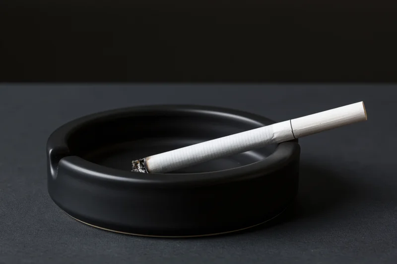 Lit cigarette resting in notch of black round ashtray on dark surface