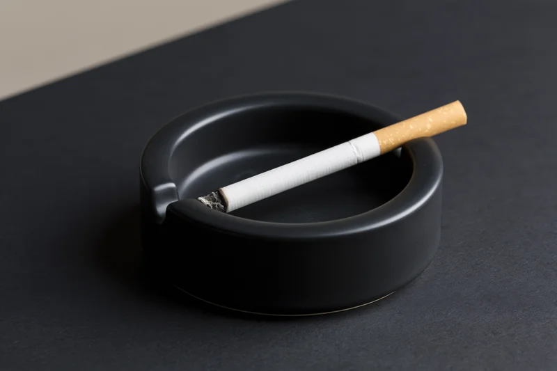 Lit cigarette resting in notch of black round ashtray on dark surface