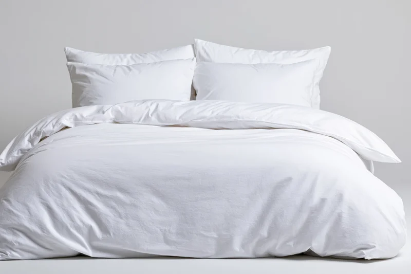 Neatly made bed with white comforter and four pillows against plain white background
