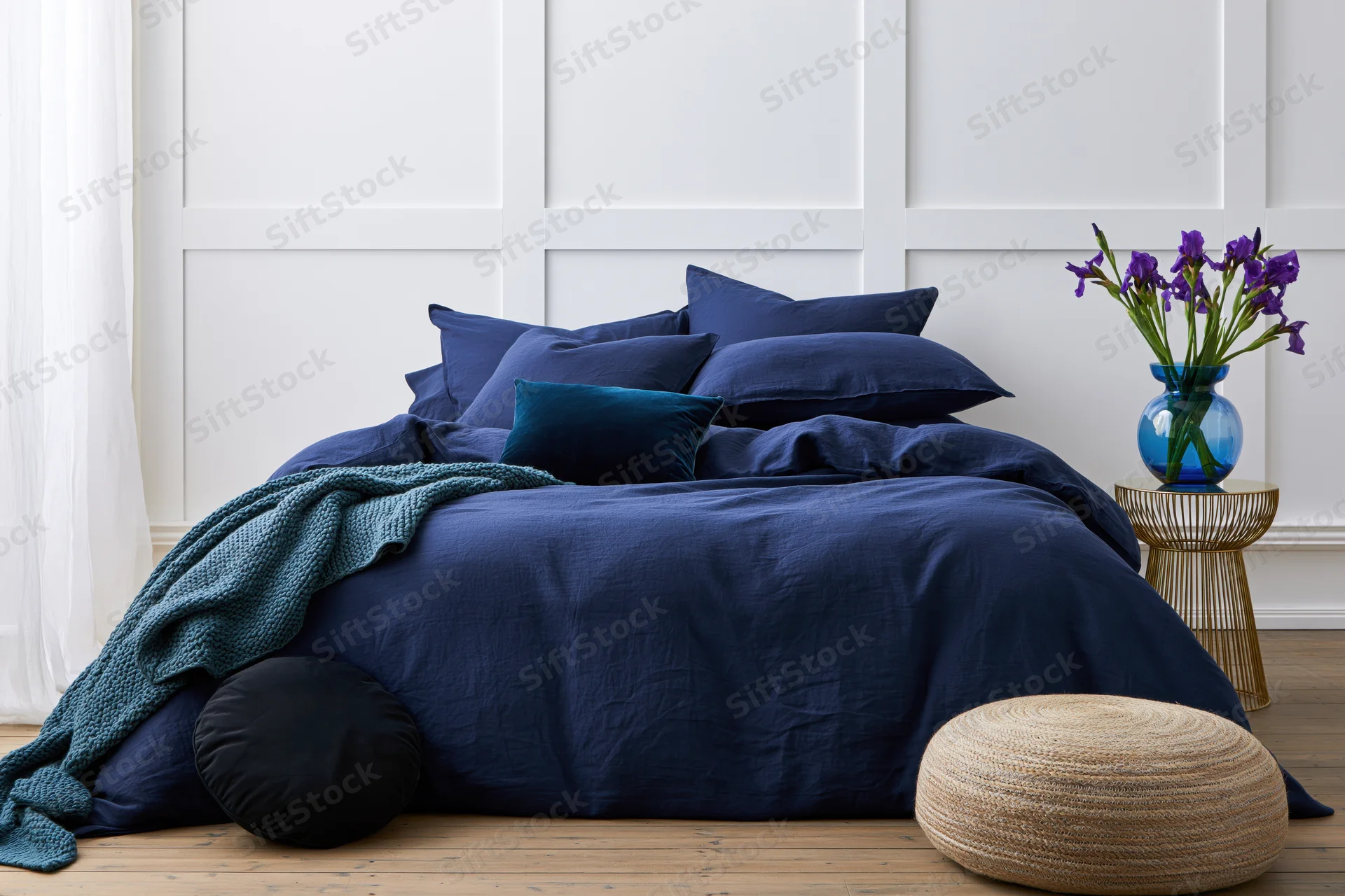 Neatly made double bed with navy blue bedding and pillows in a white paneled bedroom with a gold side table holding purple irises and a woven ottoman on wooden floor