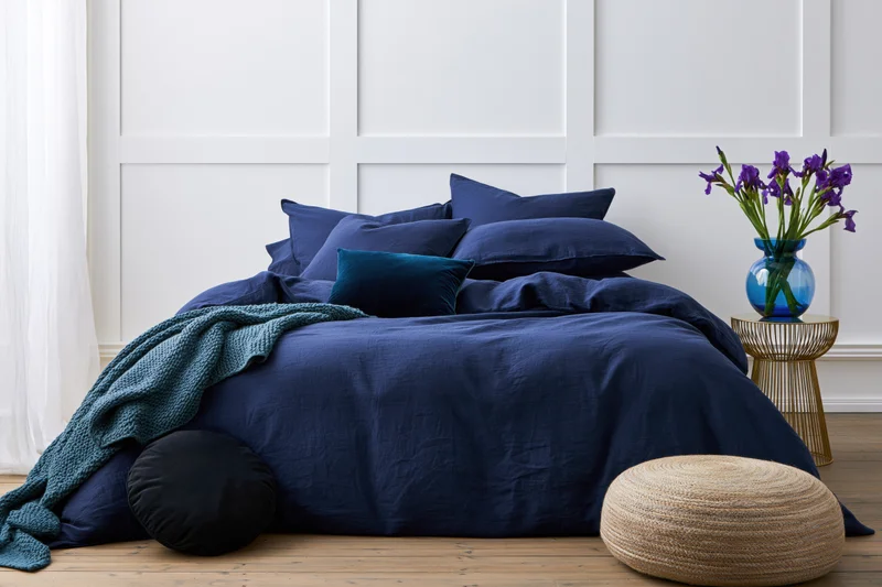 Neatly made double bed with navy blue bedding and pillows in a white paneled bedroom with a gold side table holding purple irises and a woven ottoman on wooden floor
