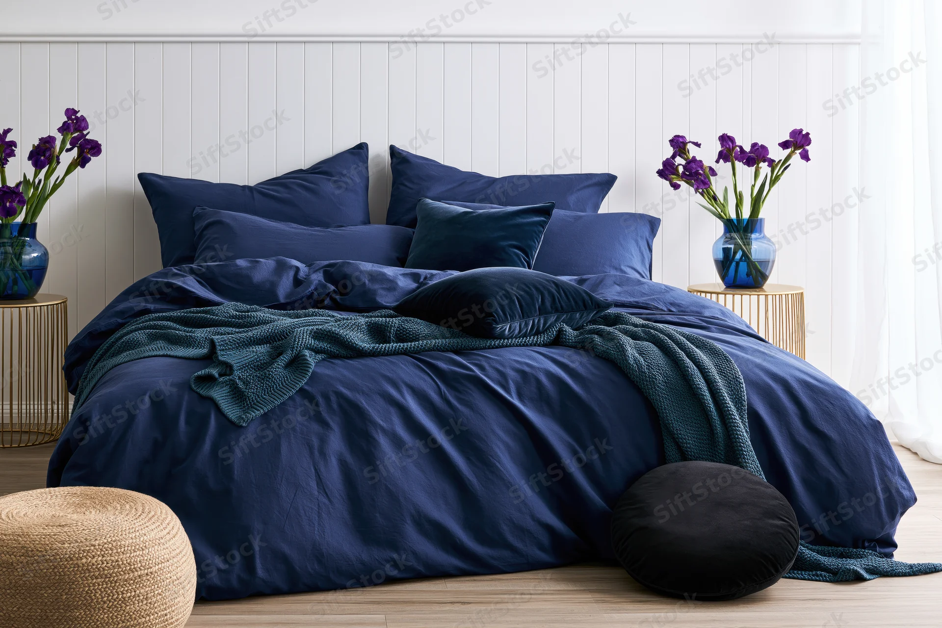 Navy blue bedding arranged on a double bed with purple irises in vases on side tables in a white paneled bedroom