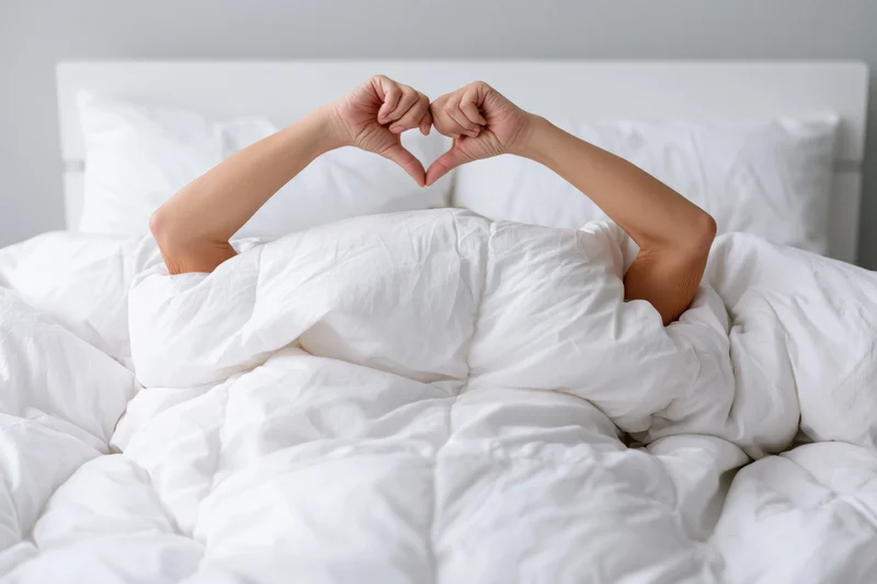 Woman under white comforter making heart shape with arms in bed