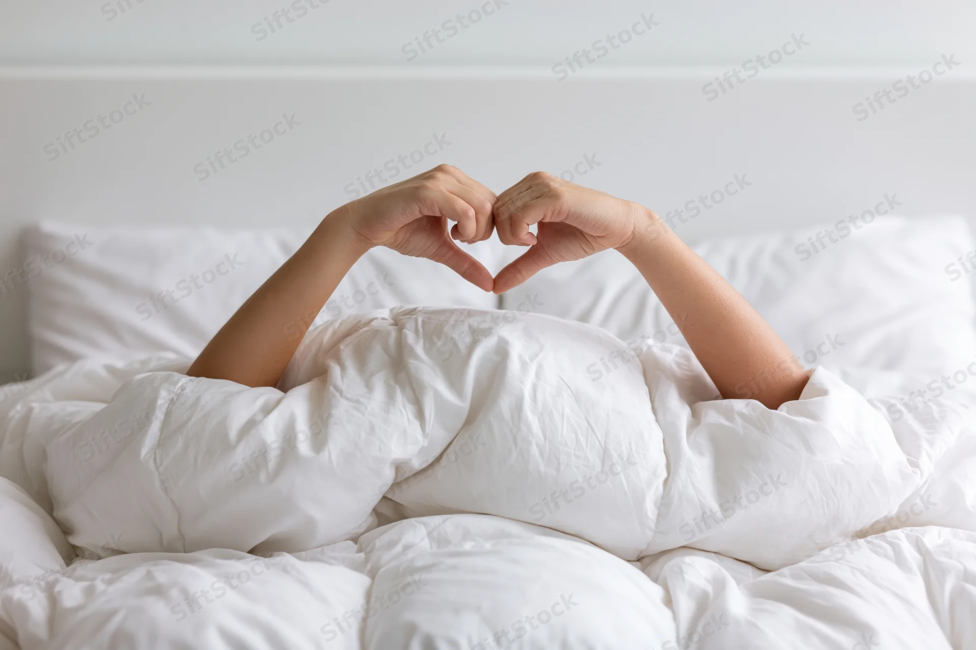 Woman under white comforter making heart shape with hands in bed