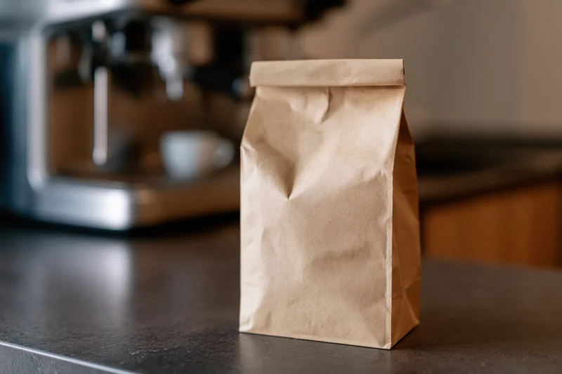 Kraft paper coffee bag standing upright on dark kitchen countertop near espresso machine