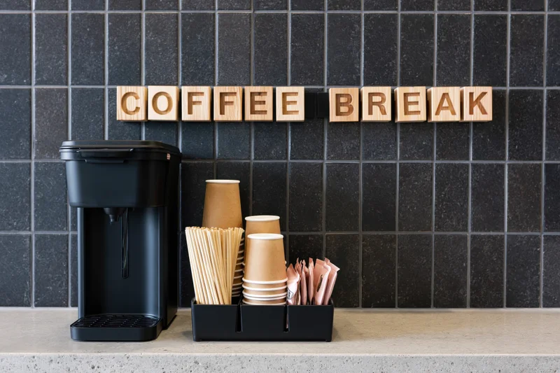 Self-serve coffee station with coffee maker and supplies on a concrete counter against black tiled wall