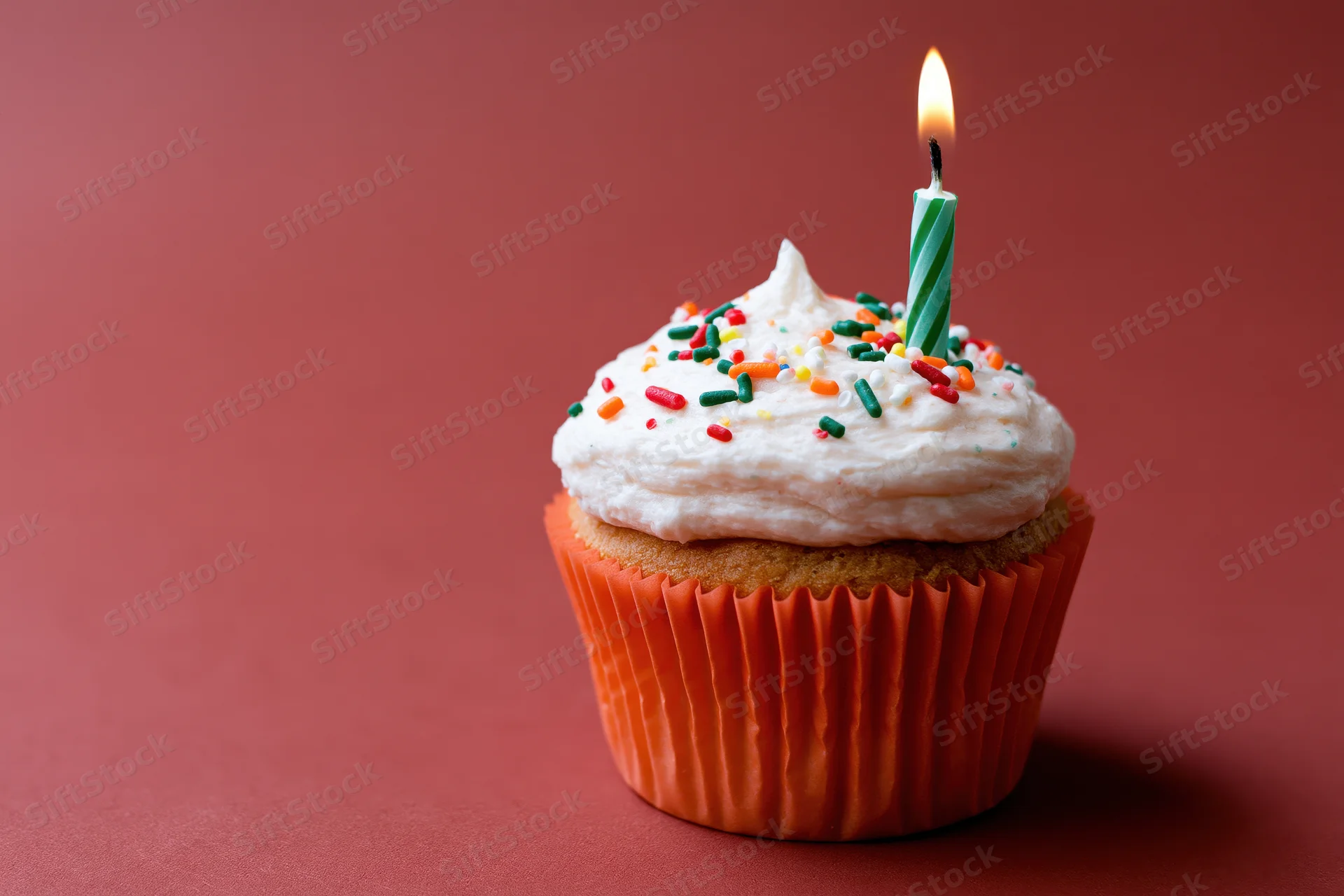 Single birthday cupcake with lit candle and sprinkles in orange paper liner on red background
