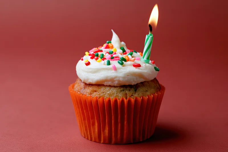 Single birthday cupcake with lit candle and sprinkles in orange paper liner on red background