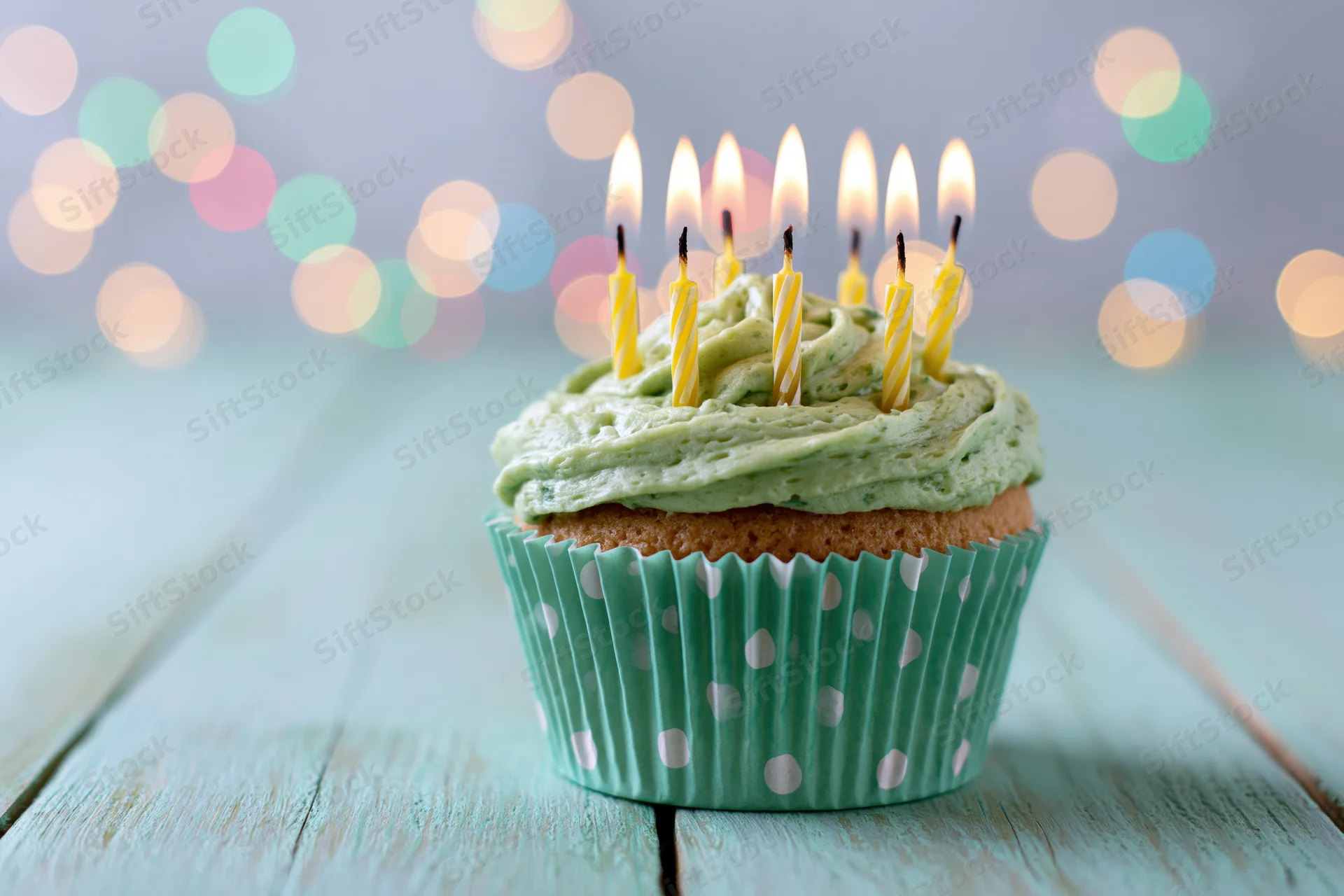 Single birthday cupcake with green frosting and six lit yellow candles on a wooden table with bokeh lights in the background