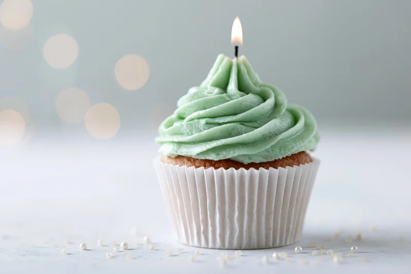 Single birthday cupcake with mint green swirled frosting and lit candle on white surface with bokeh background