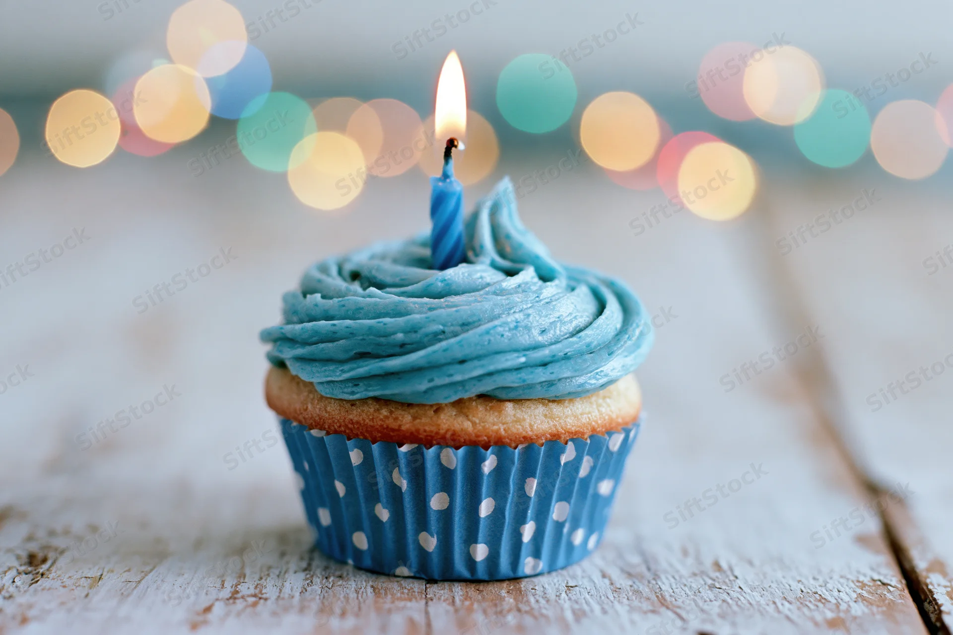 Single birthday cupcake with blue swirled frosting and one lit candle on wooden table with bokeh lights behind