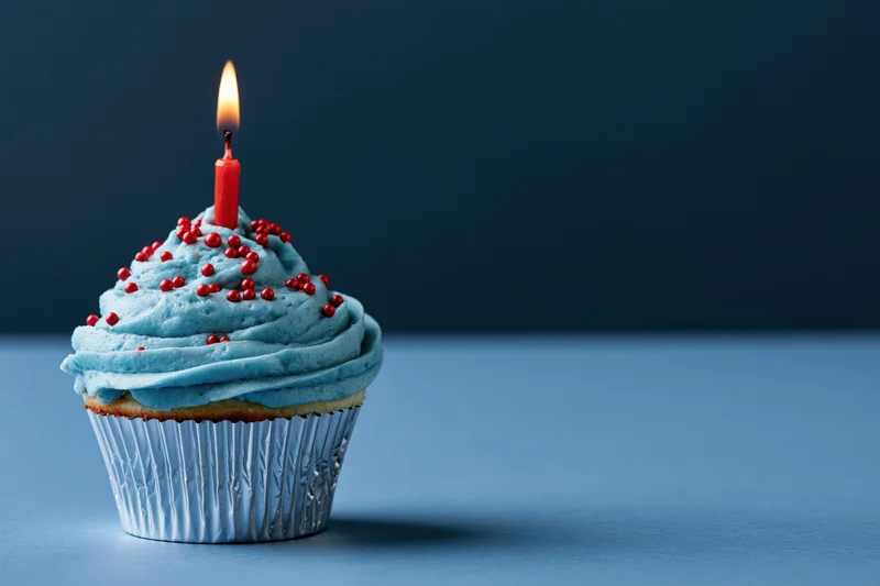 Single birthday cupcake with blue frosting and red sprinkles, lit candle, on blue surface