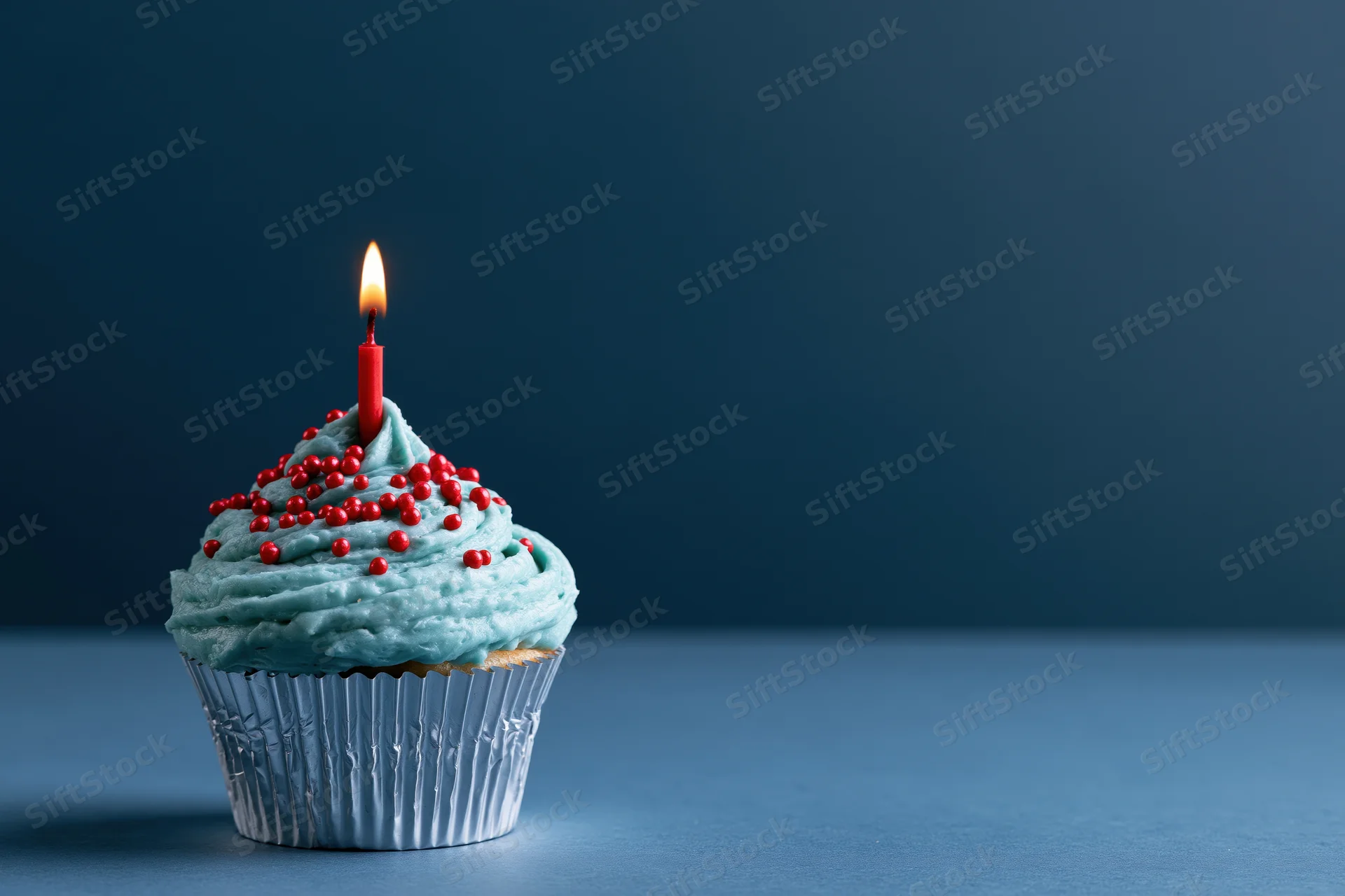 Single birthday cupcake with blue frosting and red sprinkles, lit candle, on blue surface