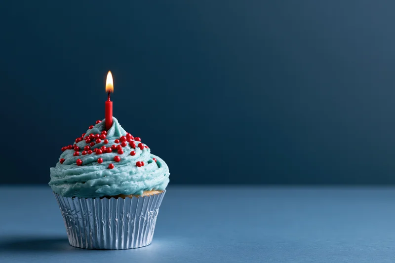 Single birthday cupcake with blue frosting and red sprinkles, lit candle, on blue surface