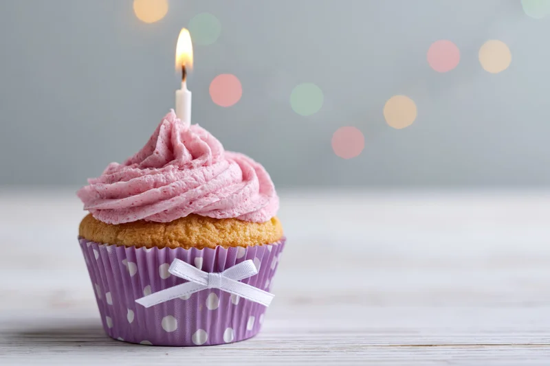 Single birthday cupcake with pink frosting and lit candle in purple polka dot liner on wooden surface